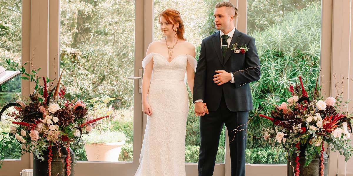 Exquisite Ceremony Flowers A smiling bride and groom at a wedding ceremony at Trevenna Barns in Cornwall, flanked by milk churns with exquisite ceremony flowers designed and created by wedding florist in Plymouth, Artem Florum.